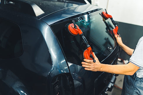 A mechanic installs a rear windshield on a dark vehicle using red suction tools.