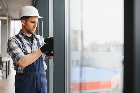A construction worker in overalls and a hard hat uses a power drill on a window frame inside