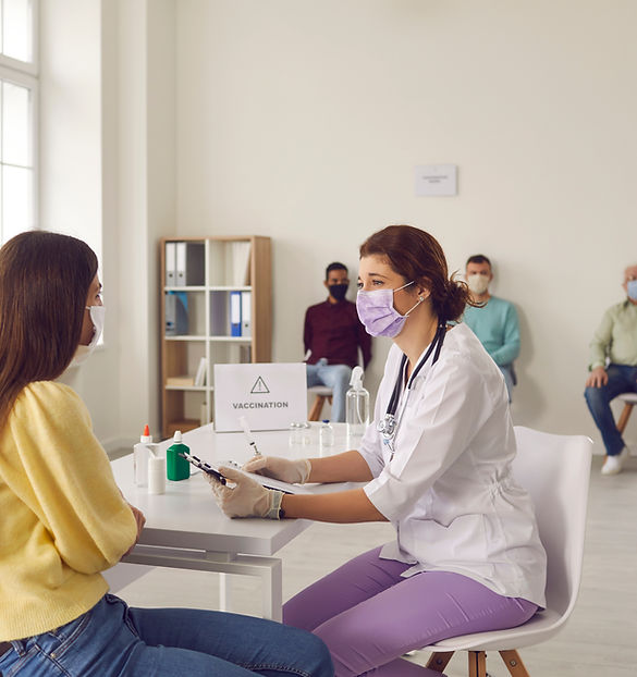A masked nurse in a white coat and gloves sits at a table with a woman in a yellow sweater