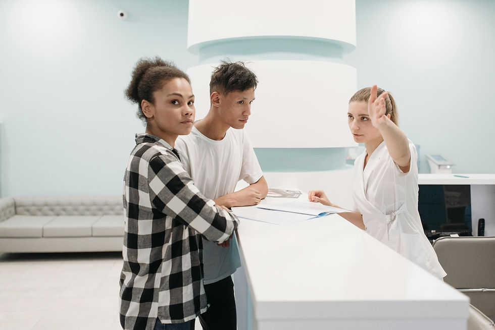 Two people waiting at a reception desk, a woman in a white uniform is pointing her hand out to the left, looking directly at the camera, and saying, "Next."