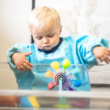 A young child in a blue smock explores a colorful spinning water wheel at the sensory table in Seward Children’s Museum, deeply focused on the hands-on activity.