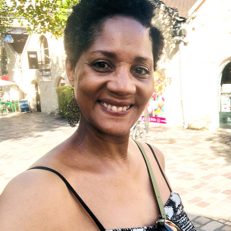 Femme souriante en robe noire et blanche dans une rue ensoleillée, sous des arbres, devant des façades claires.