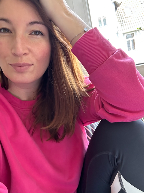 Je souris à la caméra, je porte mes cheveux lâchés, un pull rose et des leggings bleu foncé.