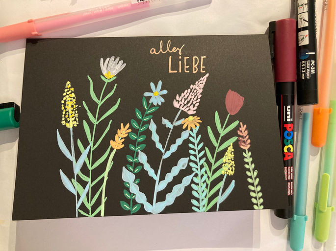 Das Bild zeigt eine schwarze Karte mit farbenfrohen Zeichnungen von Blumen und Pflanzen. In der Mitte der Karte ist der handgeschriebene Text „alles LIEBE“ in einer Mischung aus eleganter Kursivschrift und Großbuchstaben. Die Blumen sind in lebhaften Farben wie Gelb, Weiß, Blau, Pink und Rot gemalt, ergänzt durch grüne Stiele und Blätter. Um die Karte herum liegen verschiedene Marker und Stifte, darunter ein pinkfarbener Stift, ein schwarzer Posca-Marker und ein türkisfarbener Stift.
