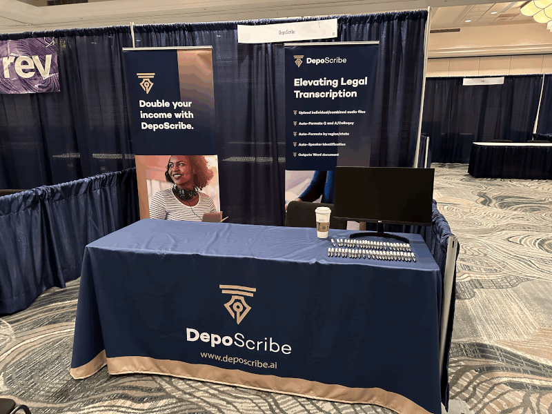 A picture of the DepoScribe booth at the AAERT Annual Conference in St. Louis, Missouri. The booth is a table with a drop cloth showing the DepoScribe logo and web address. On the table is a monitor and pens laid out for attendees. Behind the table are two standup banners for DepoScribe with the taglines "Double your income with DepoScribe" and "Elevating Legal Transcription".
