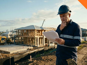 A construction worker wearing a dark blue hard hat and navy work shirt with reflective strips stands on a residential building site, thoughtfully reading a document while touching his chin. Behind him, timber-framed houses under construction, scaffolding, a concrete slab, and construction equipment are visible. An orange banner in the top-right corner reads "QLD."