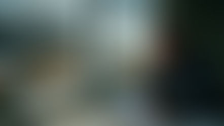 A worried construction worker sits by a window holding paperwork, looking out over a large construction site with cranes, excavators, and a bridge under a cloudy sky.