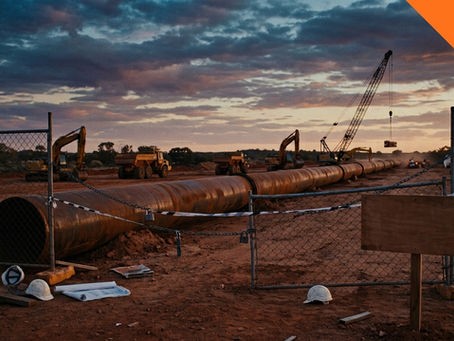 A wide-angle photo of a large pipeline construction site at dusk or dawn. In the foreground, white hard hats and blueprints rest on red dirt near a chain-link fence. The fence has caution tape and a blank wooden sign. Behind it, a long, rusty pipeline trench extends, with multiple yellow excavators, dump trucks, and a crane active under a dramatic, cloudy sky. A prominent orange banner in the top-right corner contains the text "QLD".