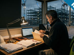 A candid photograph shows a middle-aged man with grey hair, wearing a dark fleece and work pants, seated at a cluttered wooden desk in a dimly lit site office at night. He is hunched over, reviewing documents in an open folder, illuminated by a desk lamp. On the desk are stacks of paper, rolled plans, a laptop, and a white hard hat. Through large, rain-streaked windows behind him, a brightly lit construction site with two tower cranes and a steel frame building is visible against the dark, cloudy sky.