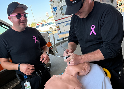 Two EMTs with pink ribbons train on a medical dummy