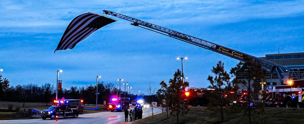 American flag draped from Pierce Manufacturing fire truck ladder, emergency vehicles at dusk.
