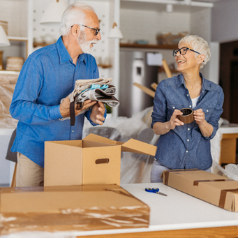 Smiling older couple packs moving boxes in a bright home, with tape, scissors and folded clothes on the table.