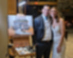 The bride and groom smiling next to their completed live painting featuring their first kiss in front of a fireplace. The painting is displayed on the artist's easel.
