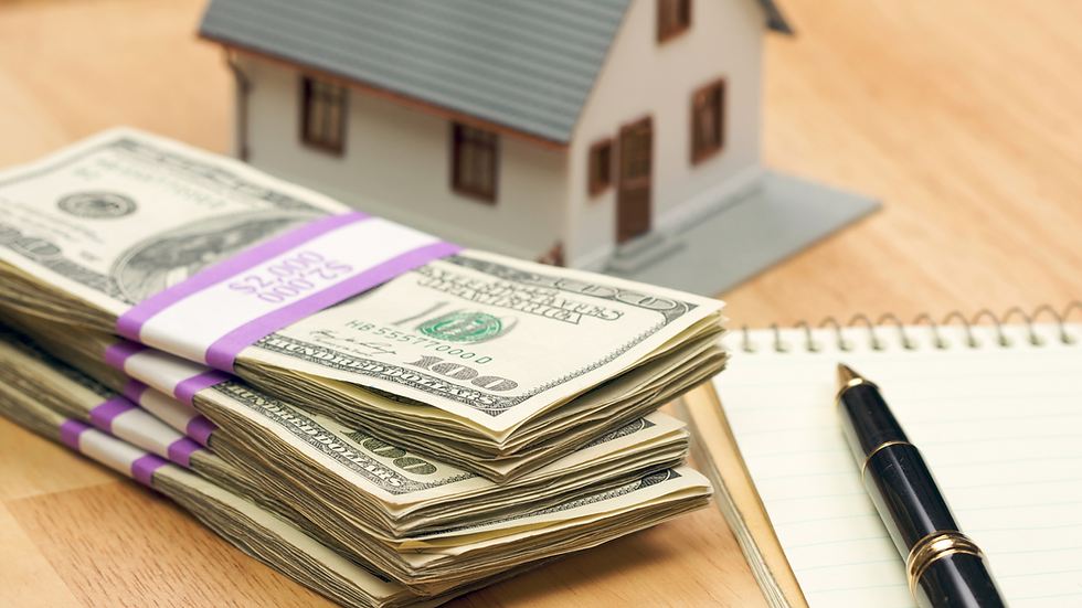 Stacks of US dollar bills, a pen, and a notepad on a table with a small model house in the background, symbolizing mortgage payoff and investment decisions.