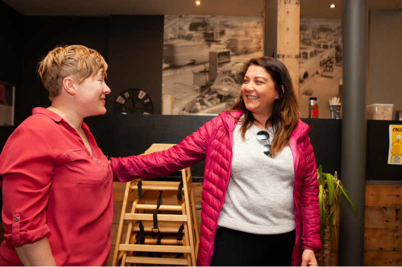 Two women, one white British with short blonde hair and the other of mixed ethnicity with long brown hair, greet each other warmly inside a coffee shop.