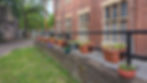 Colorful flower pots on a black railing line a brick building with arched windows. Greenery and a tree are visible in the background.