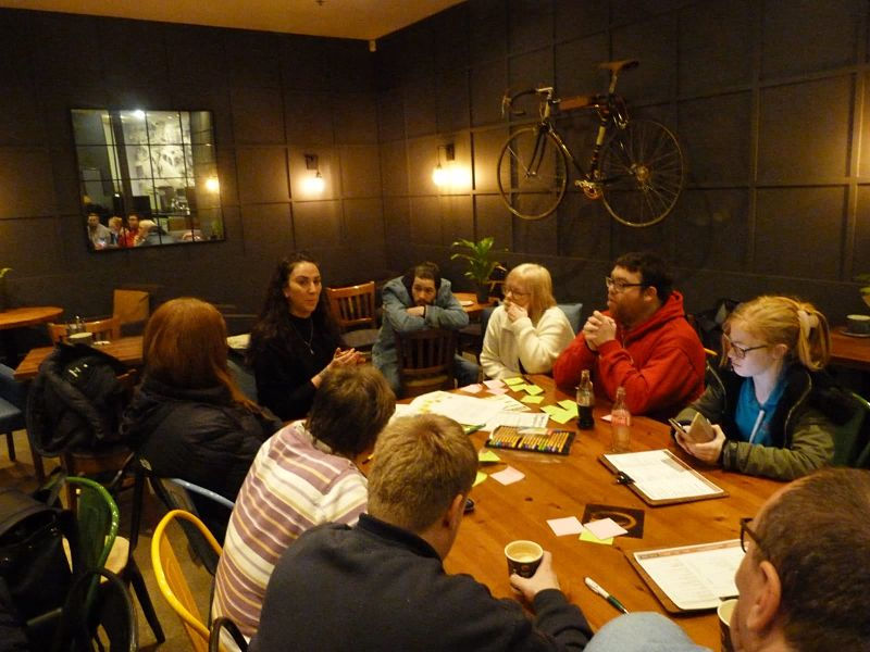 A woman with long dark hair (Melissa Smith) talks to a group sitting around a large wooden table in a coffee shop.
