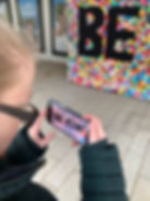 Person in a dark jacket photographs a colourful mural MADE BY Creative Kindness with "BE KIND" using a phone. The phone screen shows "BE KIND." 