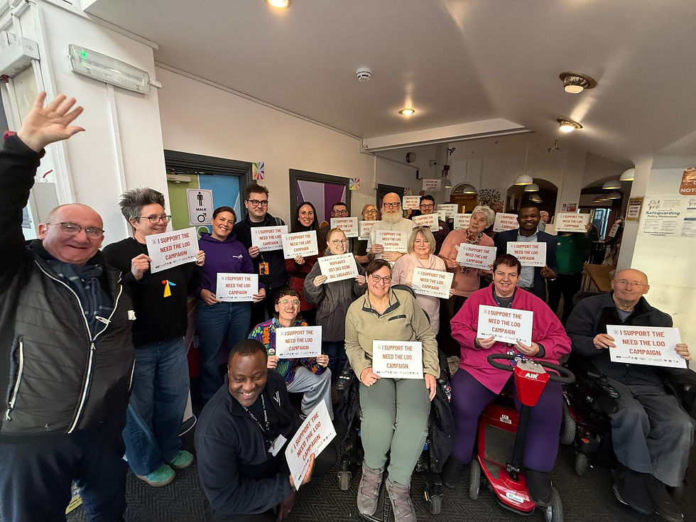 Group of smiling people indoors holding signs reading "I support the Need the Loo campaign," expressing support and enthusiasm.