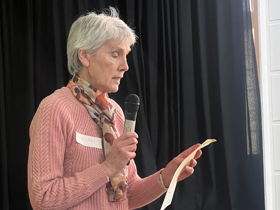 A woman in a pink sweater speaks into a microphone, holding a paper. She wears a floral scarf. Black curtains form the background.