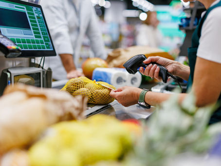 Grocery cashier scans a yellow mesh bag of potatoes at a checkout counter, with a green screen interface and shoppers blurred behind.