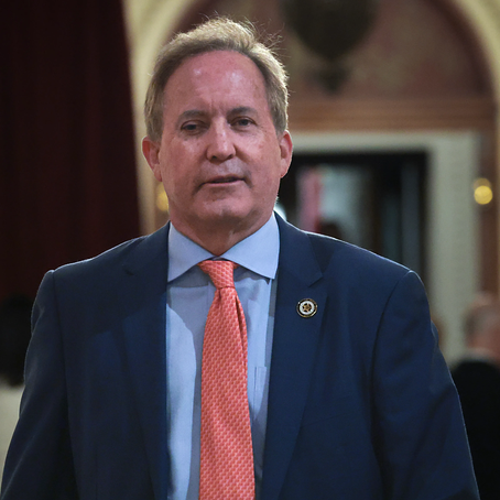 Ken paxton stands indoors, facing camera with a serious expression; blurred warm background.