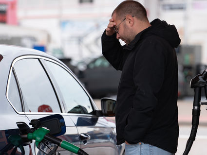 Stressed man in black jacket refuels a silver car at a gas station, hand on forehead, with fuel pump and blurry background.