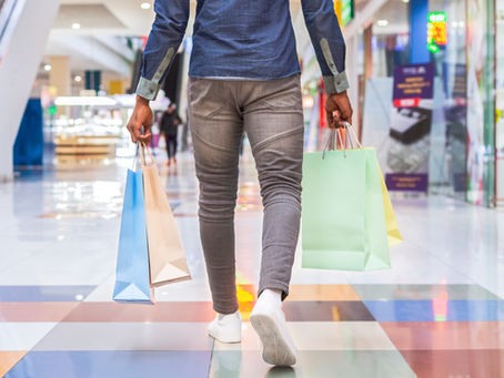 Person walking through a bright mall with colorful shopping bags, seen from behind, with blurred stores and escalators.