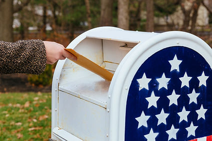 Hand inserts yellow envelope into white mailbox with blue star panel, outdoors in a park-like setting.