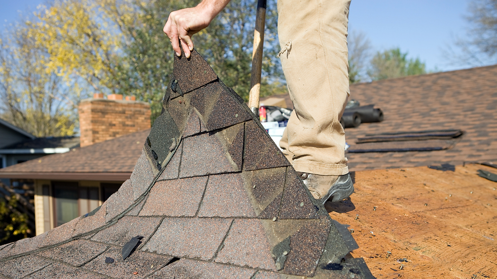 Worker removing roof shingles with a tool, set against a house and trees. The shingles are dark and patterned, conveying a focused effort.