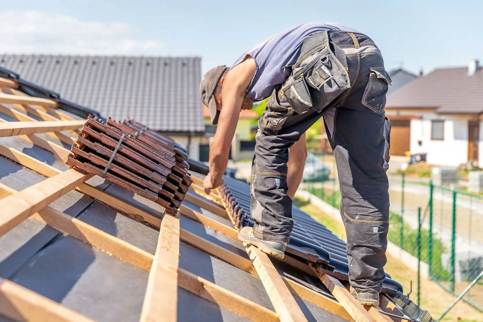 Worker on a sloped roof laying tiles under a clear sky. Wearing a cap and tool belt, with houses in the background.