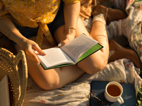 Person in yellow floral top reading a book outdoors on a blanket, with a wicker basket and cup of tea nearby. Relaxed, sunny setting.
