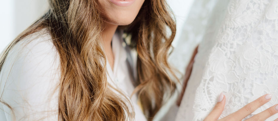 Smiling woman with long hair, wearing white, touches a lace dress. Soft natural light creates a serene, joyful mood.