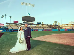 Bride in a white gown and groom in a suit stand smiling on a baseball field. Stadium seats and signs for Spectrum in the background.
