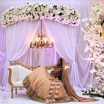 Bride in ornate attire reclines on a gold-trimmed sofa under a floral arch and chandelier, with white drapes and flowers adorning the setting.