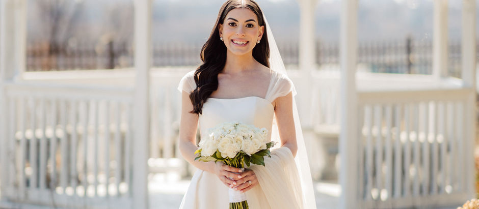 Bride in white dress holding bouquet, standing in front of a gazebo. Sunlit setting with green lawn and clear sky in the background.