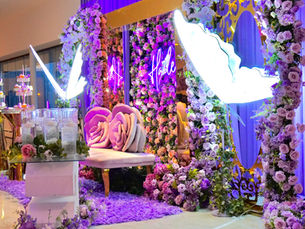 Elegant floral wedding backdrop with purple flowers, neon "Hafee" sign, a butterfly-shaped light, and plush rose chairs, exuding romance.