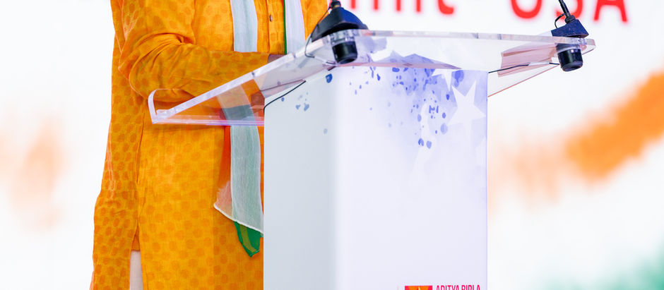 Man in an orange kurta speaks at a podium with Aditya Birla Capital logo. Background features "International Summit - USA."