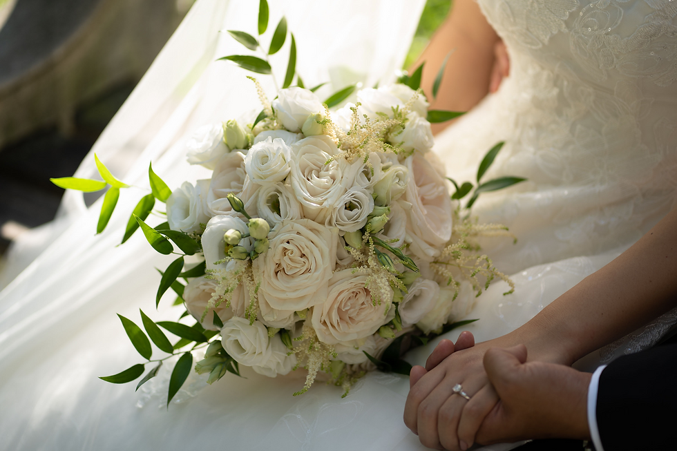 Bride holding groom's hand, displaying ring. White rose bouquet with greenery on lace dress. Sunlit romantic setting.