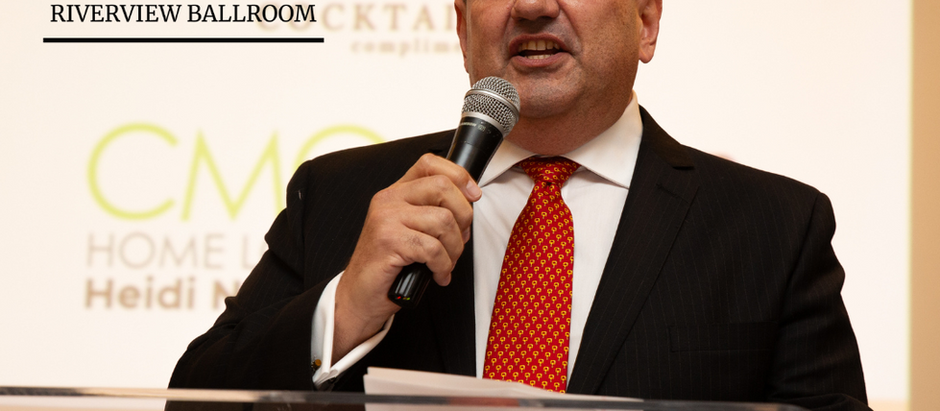 Man in a suit speaks into a microphone at a podium with "Riverview Ballroom" logo. Background shows a screen with text and a bridge.