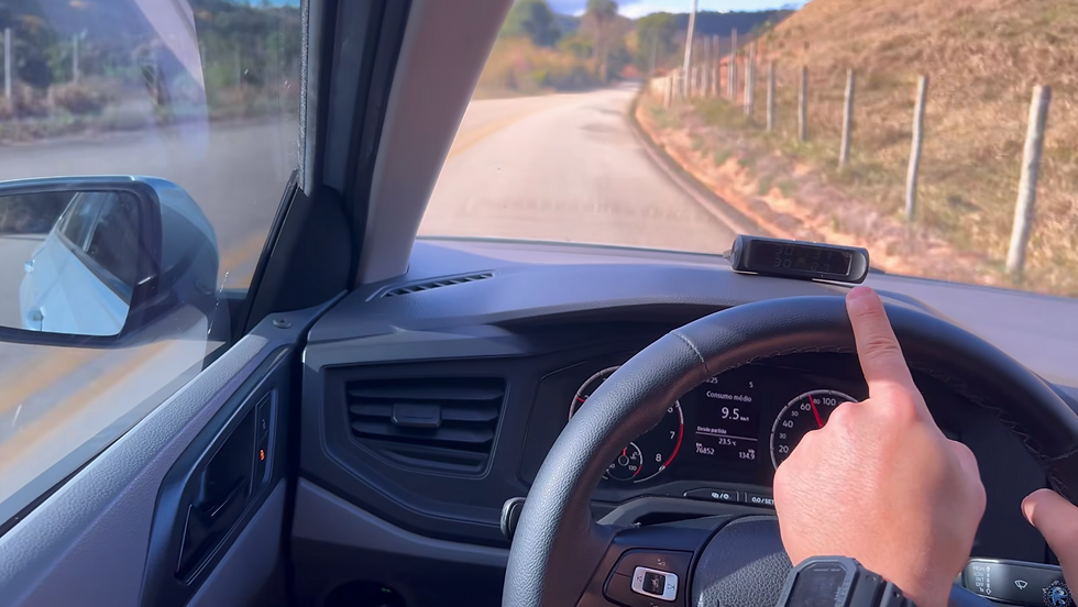 De dentro do carro, mão aponta para dispositivo no painel enquanto segue em estrada rural sinuosa; no painel, Consumo médio.