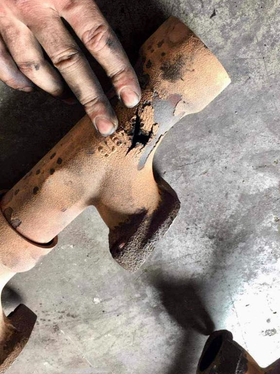 Hands point to a rusty, cracked SCANIA V8 manifold on a concrete floor, highlighting damage. The mood suggests concern .