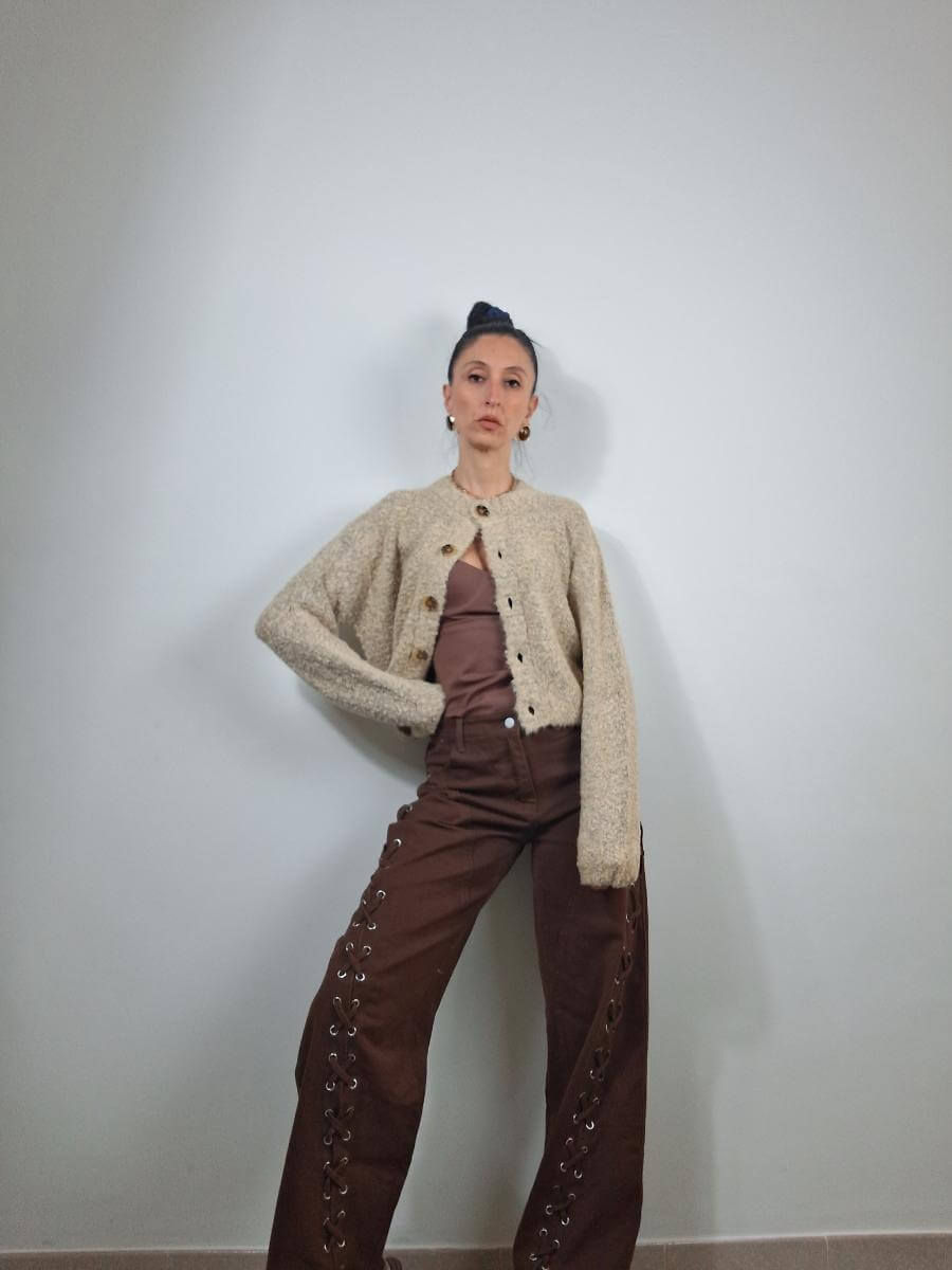 Giovane donna indossa cardigan beige e pantaloni marroni, in posa su sfondo bianco.