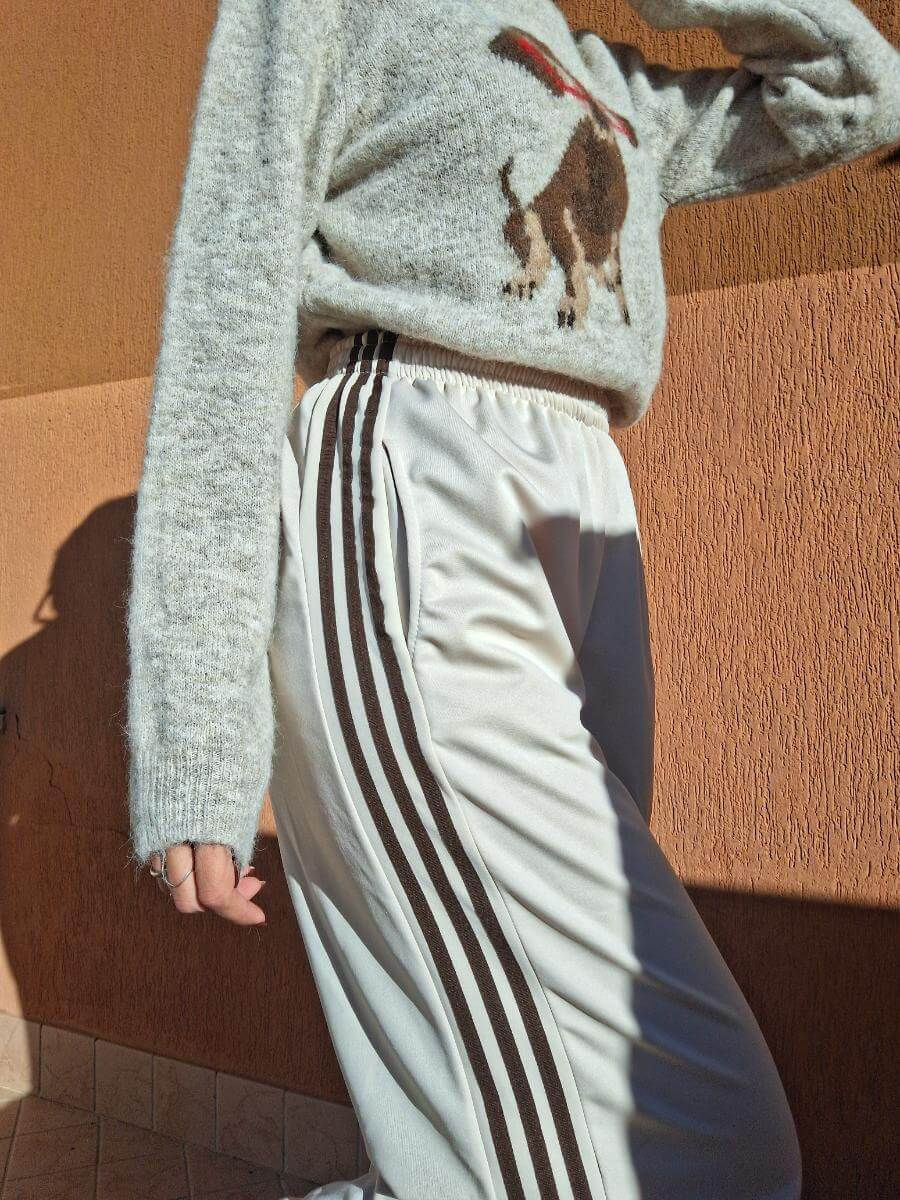 Persona che indossa pantaloni bianchi con tre strisce e maglione grigio, vicino al muro.