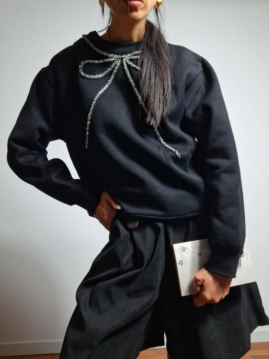 Woman wearing black sweatshirt with bow, holding a book, dark skirt, pose.