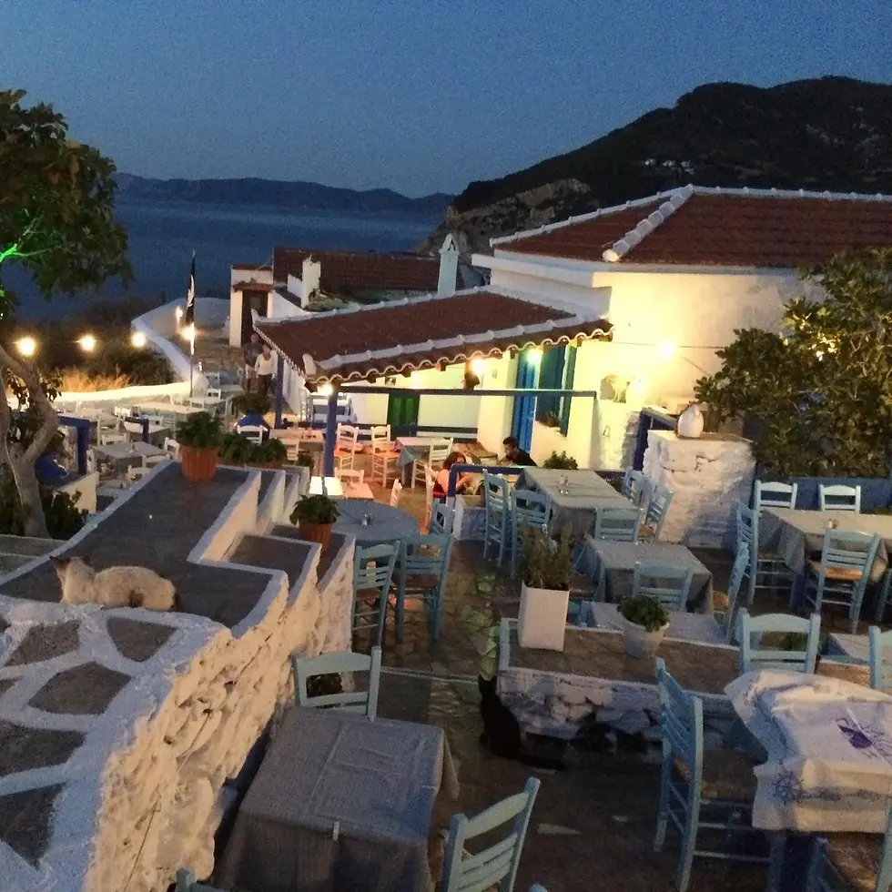 Outdoor Greek restaurant with blue chairs and white tables at dusk, overlooking a calm sea. Cozy lights illuminate the rustic setting. Greek live music
