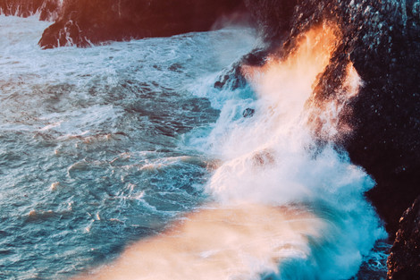 Blue waves with orange sunset glow mist crashing into a cliff side