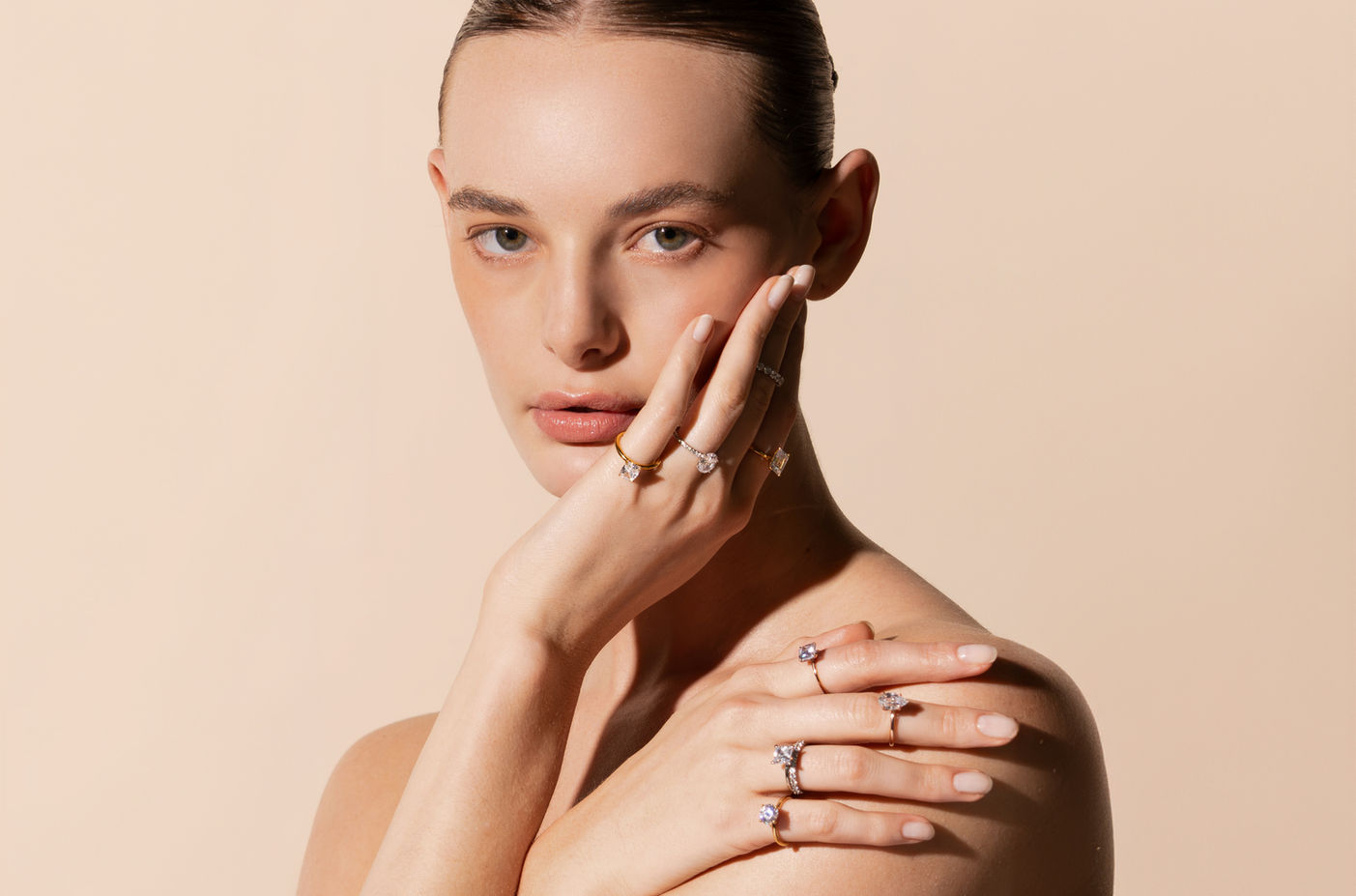 Primer plano de una modelo luciendo anillos de diamantes de Eonian Diamonds. La imagen muestra las manos de la modelo tocando delicadamente su rostro, exhibiendo múltiples anillos de compromiso y alianzas en sus dedos. Los anillos presentan diseños elegantes con diamantes cultivados en laboratorio. La modelo tiene una apariencia natural y refinada, con un maquillaje sutil, sobre un fondo de color beige claro que complementa la estética minimalista y sofisticada de la joyería.