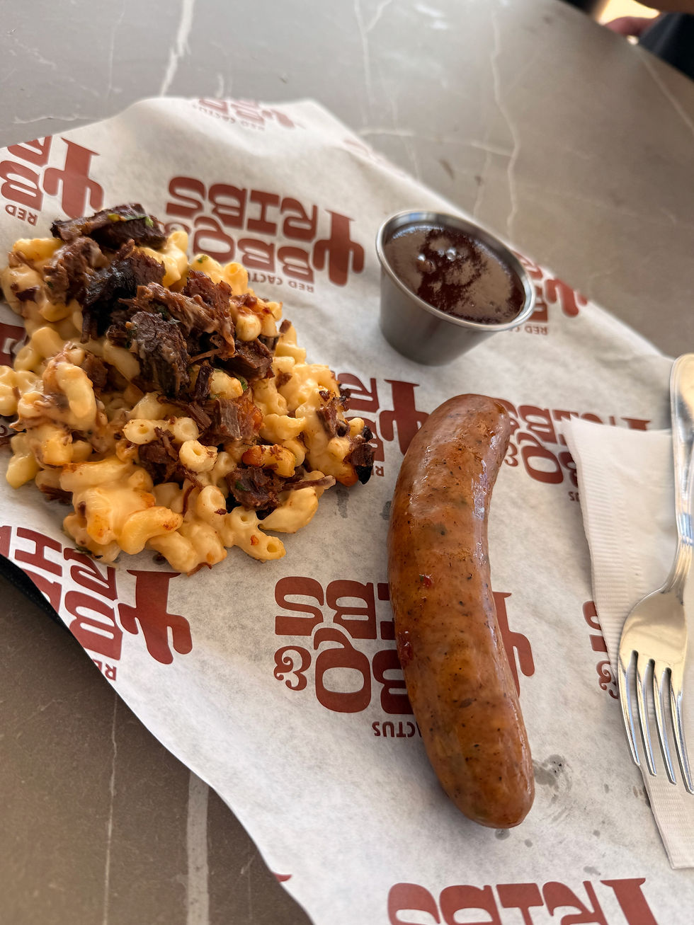 Mac and cheese topped with pulled beef, a sausage, and sauce cup on Red Hook BBQ paper at a restaurant table.