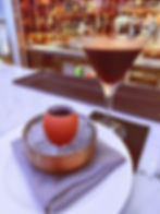 Cocktail glass with a frothy brown drink beside a cup in an eggshell on ice. Marble bar with colorful bottle display in the background.
